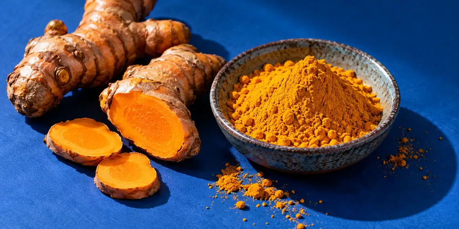Racines de curcuma frais coupées à côté d’une coupelle de poudre de curcuma orange sur un fond bleu vif