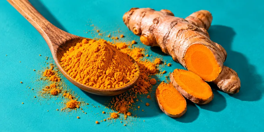 Cuillère en bois remplie de poudre de curcuma posée à côté d’un rhizome frais sur un fond turquoise vif