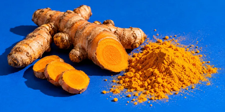 Rhizomes de curcuma frais, dont un coupé en deux, à côté d’un petit tas de poudre orange sur un fond bleu roi
