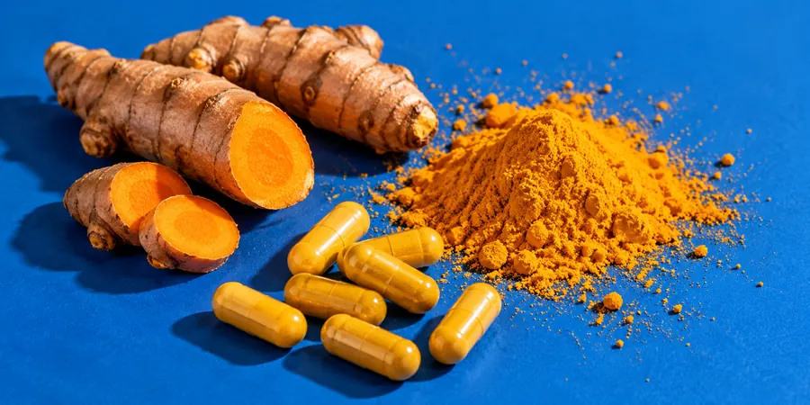 Gélules jaunes sans marque éparpillées autour d’une racine de curcuma fraîche et d’un petit tas de poudre orangée, sur un fond bleu vif
