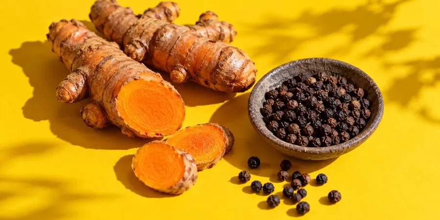 Racines de curcuma fraîches, dont une coupée en deux, et grains de poivre noir dans une coupelle en céramique sur un fond jaune vif
